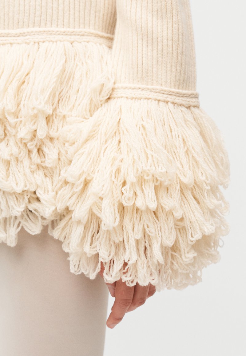 Suéter de punto con mangas grandes y esponjosas con flecos color marfil y cuerpo acanalado. Presenta un color sólido en contraste en el puño y una textura suave.