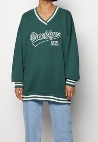 Sweatshirt vert surdimensionné avec un graphisme blanc Brooklyn et N.Y. ; possède un col en V, des poignets rayés et un ourlet.