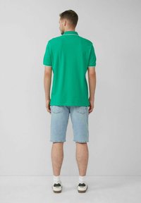 Grünes Poloshirt aus Baumwolle, mit kurzen Ärmeln und Kragen. Kombiniert mit hellen Jeansshorts und weißen Sneakers vor einem neutralen Hintergrund.