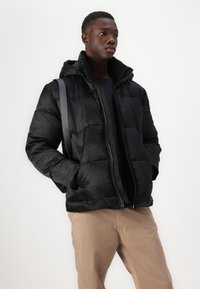 Melna pūka jaka ar cepuri, ar izsmalcinātu logo rakstu, ar rāvējslēdzēju un sānu kabatām, uzvilkta virs melna krekla un bēšiem biksēm.