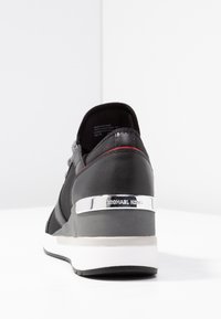 Czarne sneakerki z białą podeszwą i srebrnym akcentem na pięcie z grawerem "Michael Kors" na jednolitym białym tle.