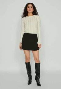 White long-sleeve top with vertical ribbing, paired with a black mini skirt. Model wears knee-high black boots. Simple, fitted design.