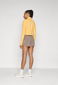 Idrottskläder med en gul cropped långärmad topp och bruna shorts med rosa detaljer; mönstrad sömnadsdetaljer synliga.