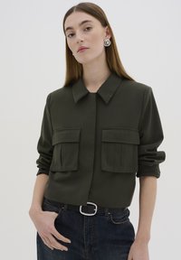 Chemise coll à col vert foncé avec deux poches poitrine, fermeture à boutons, manches retroussées et tissu lisse. Portée avec un jean en denim foncé taille haute et une ceinture.