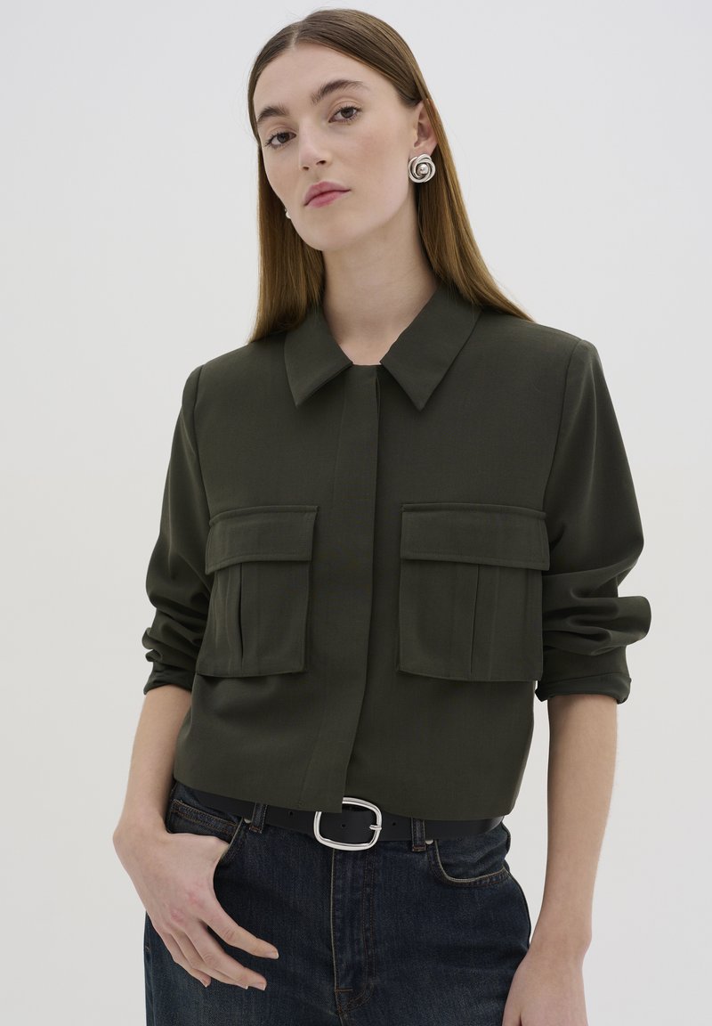 Chemise coll à col vert foncé avec deux poches poitrine, fermeture à boutons, manches retroussées et tissu lisse. Portée avec un jean en denim foncé taille haute et une ceinture.