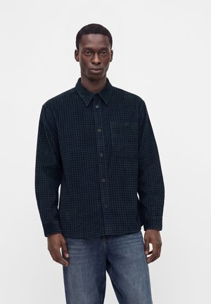 Jeune homme portant une chemise à carreaux bleu foncé à manches longues et un jean bleu, debout devant un fond clair uni.
