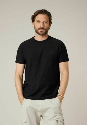 Man met baard, gekleed in een zwart T-shirt met korte mouwen en lichtgekleurde cargobroek, staand met handen in de zakken tegen een effen achtergrond.