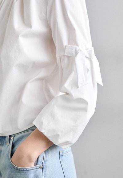 Fehér pamut blúz, puffos ujjal, a mandzsettákon megkötő részletekkel. Világoskék farmernadrággal párosítva, amely laza szabású.