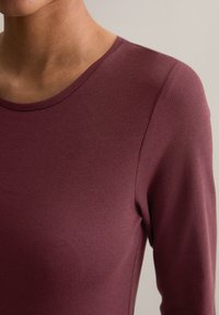 Bordeaux langermet topp laget av ribbet stoff; har rund halslinning og tettsittende design. Glatt tekstur med minimale sømmer.