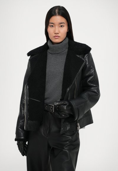 Črna usnjena jakna z strukturiranim črnim ovčjim krznom na ovratniku, dolgimi rokavi in srebrnimi zadrgami, v kombinaciji s sivo turtleneck majico.