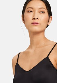 Camisola nera con spalline sottili, scollatura a V, bordi ondulati e una texture liscia e morbida. Il tessuto sembra leggero e elasticizzato.