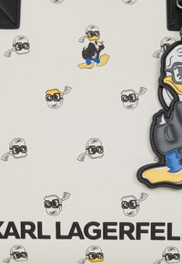 Witte tas met herhaalde cartoon-eend graphics in het zwart en geel, met een prominente zwarte tekst die "KARL LAGERFELD" op de onderste rand leest.