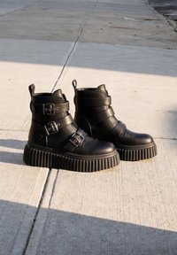 Μαύρα μποτάκια πλατφόρμας με παχιά ανάγλυφη σόλα και τρεις λουριά με πόρπες σε κάθε μποτάκι, τοποθετημένα σε ηλιόλουστο τσιμεντένιο πεζοδρόμιο.