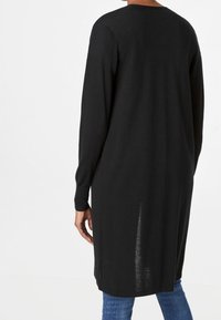 Henkilö, jolla on pitkä musta neuletakki pitkillä hihoilla, seisoo selkä katsojaan päin, yhdistettynä sinisiin farkkuihin.