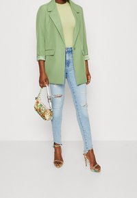 Blazer verde oversize sopra una maglietta verde chiaro, abbinati a jeans chiari strappati e borsa multicolore decorata. Sandali con tacco a listini.