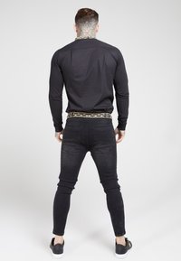 Hombre con tatuajes en el cuello y las manos, vistiendo una camiseta de manga larga negra, una cinturilla negra con patrón, jeans ajustados negros y zapatillas negras.