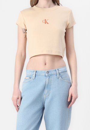Kobieta nosząca beżowy krótki t-shirt Calvin Klein i jasnoniebieskie jeansy z wysokim stanem, pokazująca tatuaż na lewej ręce.