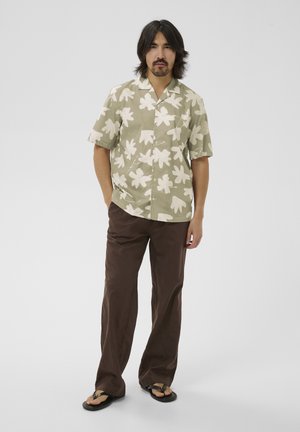 Mand stående med hænder i lommerne iført beige blomstret kortærmet skjorte, brune bukser og sorte flip-flops på en hvid baggrund.