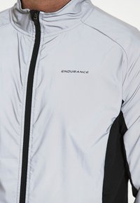 Lys grå sportsjakke med sorte accenter, lynlås fortil, høj krave, "ENDURANCE" logo på brystet og glat tekstur.