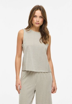 Kvinne med lyst brunt hår iført ermeløs, beige og hvit topp med vertikale striper og matchende bukser, stående mot enkel hvit bakgrunn.