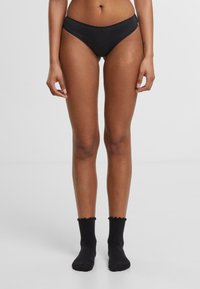 Svarte bikiniunderdeler med glatt tekstur, mid-rise design og minimalistisk stil, kombinert med svarte, ribbede sokker med scallop-kanter.