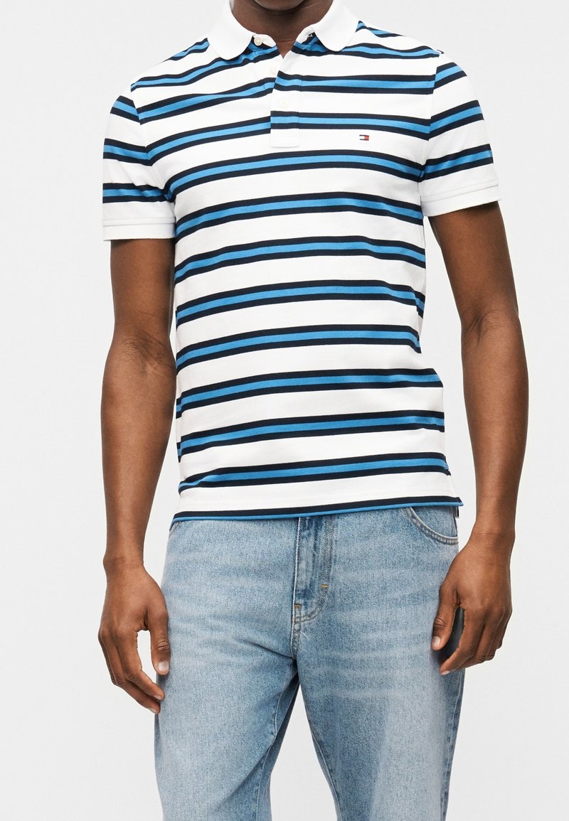 Homme portant un polo à manches courtes avec des rayures horizontales bleues et noires et un jean bleu clair sur un fond uni.