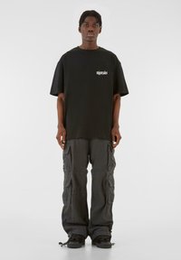 Black oversized t-shirt with a white logo in the upper left, paired with gray cargo pants featuring multiple pockets and a relaxed fit.