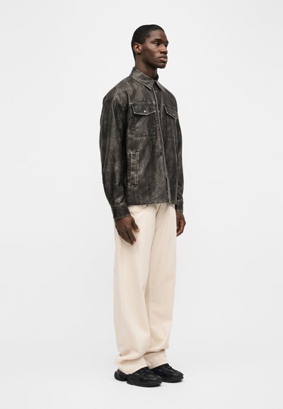 Άνδρας σε πλευρική στάση, φορώντας σκούρο καφέ δερμάτινο μπουφάν, εκρού παντελόνια φαρδιάς ραφής και μαύρα αθλητικά παπούτσια, σε απλό λευκό φόντο.