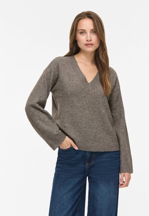 Kvinde med langt hår iført en brun V-hals sweater og blå jeans, står med den ene hånd i lommen mod en ensfarvet baggrund.