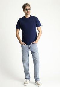 Granatowy t-shirt z krótkim rękawem, okrągły dekolt, w połączeniu z jasnoniebieskimi dżinsami i szarymi trampkami z białymi podeszwami. Zielone okrągłe okulary przeciwsłoneczne.