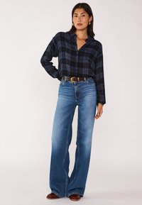 Camicia a quadri nera e blu con colletto, bottone davanti, abbinata a jeans a gamba larga blu chiaro e una cintura marrone. Stivali in camoscio marroni completano il look.