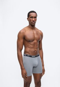 Άνδρας σε φόρμα, όρθιος, χωρίς πουκάμισο, φορώντας γκρι boxer της Nike με μαύρη ζώνη μέσης και λευκό λογότυπο, σε απλό λευκό φόντο.