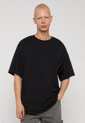 Junge Person mit kurzen blonden Haaren, trägt Ohrringe, ein schwarzes, übergroßes T-Shirt und graue Hosen, steht mit einer Hand in der Tasche.