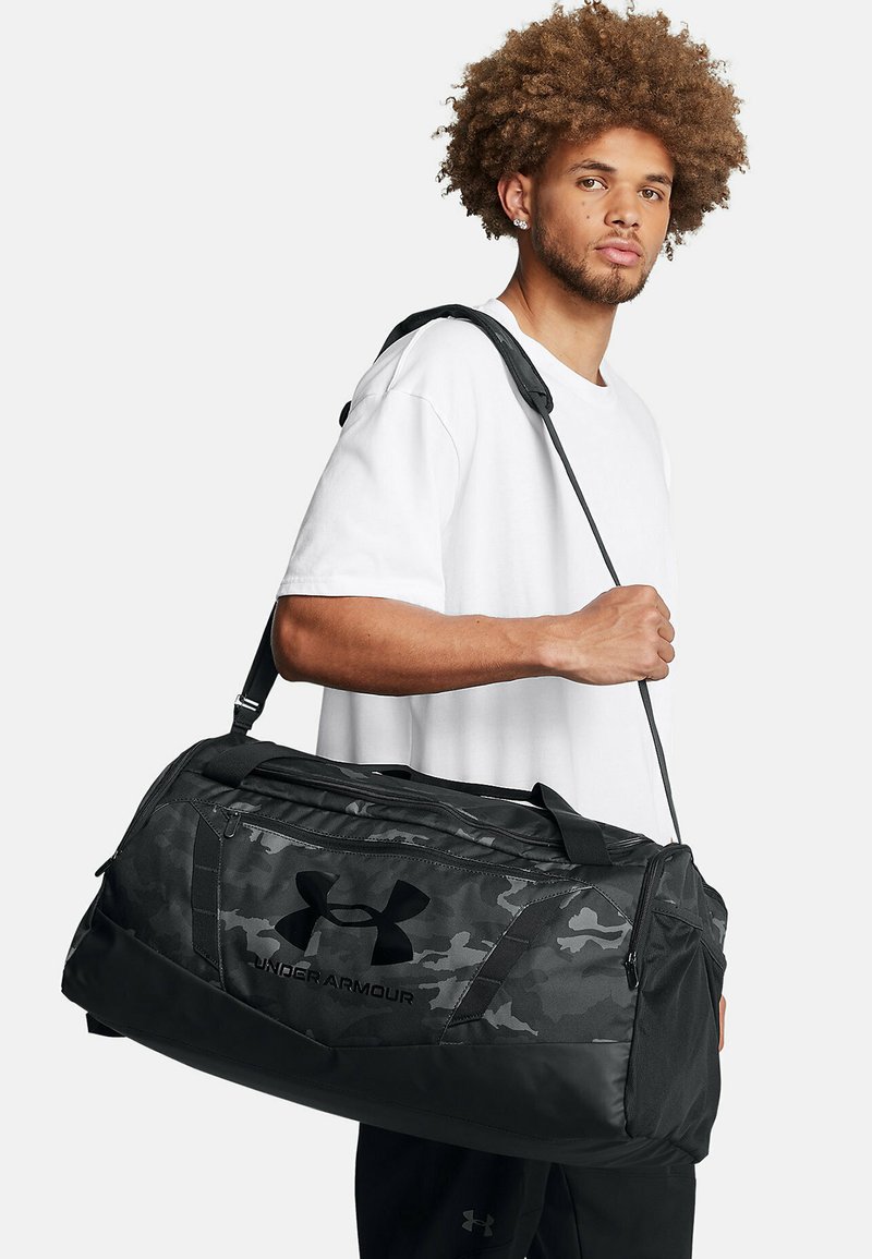 Black duffel bag with a camouflage pattern, long adjustable strap, textured fabric, and a prominent black logo on the side.