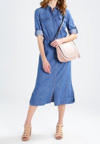 Nainen, joka on pukeutunut keskipitkään siniseen farkkuhenkilöön, beigeihin avokassyihin ja kantaa vaaleanpunaista ristivartalolaukkua, jossa on rei'itettyjä yksityiskohtia.