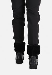 Botas negras de tobillo con puños gruesos y esponjosos, suela robusta y acabado de cuero suave. Combinadas con pantalones negros, mostrando un diseño casual y cálido.