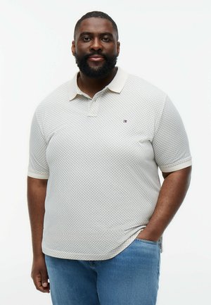 Mand med skæg iført en hvid poloshirt med mønster og blå jeans, stående med den ene hånd i lommen foran en hvid baggrund.