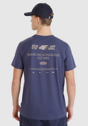 Młody mężczyzna widziany od tyłu, ubrany w granatowy t-shirt z beżowym tekstem graficznym i granatową czapkę z daszkiem.