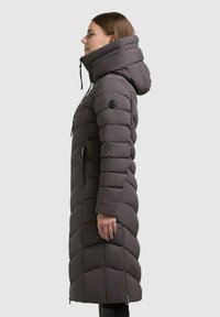 Donna di profilo che indossa un lungo cappotto invernale grigio scuro trapuntato con un alto colletto imbottito e cappuccio su uno sfondo semplice.