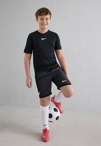 Poiss mustas Nike spordiriietuses tasakaalustab jalgpalli parema jalaga, kandes valgeid sokke ja roosaid krampe, naeratades silmad kinni.