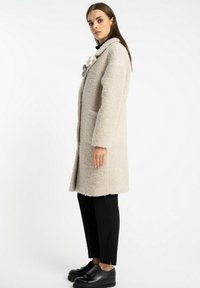 Beżowy, teksturowany płaszcz wełniany z kołnierzem typu szal i bocznymi kieszeniami. Posiada dekoracyjne czarne i szare akcenty przy dekolcie. Noszony z czarnymi spodniami.