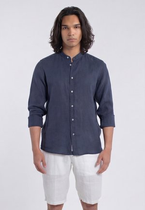 Jonge man met lang donker haar, gekleed in een marineblauw, geknoopt overhemd met lange mouwen en witte korte broek, staand tegen een effen lichte achtergrond.