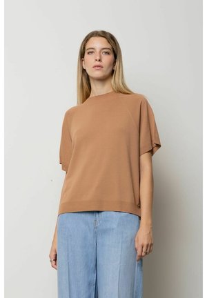 Donna con lunghi capelli biondi che indossa una maglietta corta marrone larga e jeans azzurri a vita alta, in piedi contro uno sfondo semplice.