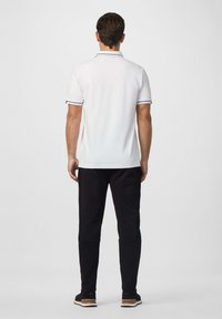 Muž stojící zády, oblečený v bílém polo tričku s krátkým rukávem a černým lemem, černých kalhotách a černých teniskách s hnědými podrážkami.