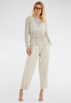 Mujer con suéter beige transparente de punto acanalado, pantalones blancos de pierna ancha y cintura alta, tacones blancos con tiras y gafas de sol con lentes tintadas en naranja, de pie con la mano en el bolsillo.