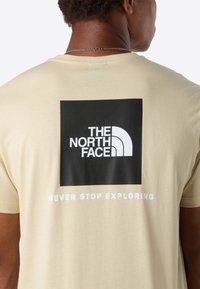 Osoba v béžovém tričku s logem The North Face a textem "Never Stop Exploring" na zadní straně, viděná zezadu.