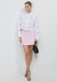 Weißes, kurz geschnittenes Hemd mit rosa gestickten X-Mustern, kombiniert mit einem rosa Miniskirt und einer schwarzen Handtasche. Schwarze, gestrickte Absätze vervollständigen das Outfit.