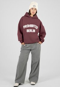 Joven mujer con un gorro de punto beige, sudadera color burdeos con el texto "PROHIBITED BERLIN", pantalones grises holgados y zapatos blancos, de pie con las manos en los bolsillos.