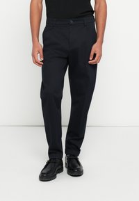 Tengerészkék egyenes szabású nadrág, övbújtatókkal és gombos rögzítéssel, fekete cipővel párosítva. Sima szövet textúra.