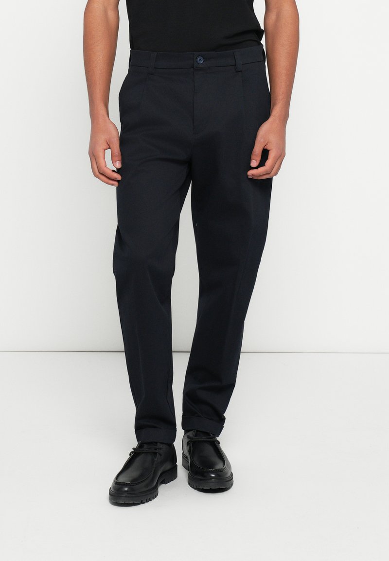 Tengerészkék egyenes szabású nadrág, övbújtatókkal és gombos rögzítéssel, fekete cipővel párosítva. Sima szövet textúra.